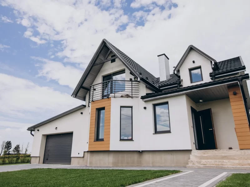 Casa moderna de dos pisos con fachada blanca y detalles en madera, ubicada en un entorno natural. Incluye un garaje y un jardín bien cuidado. Cielo parcialmente nublado.